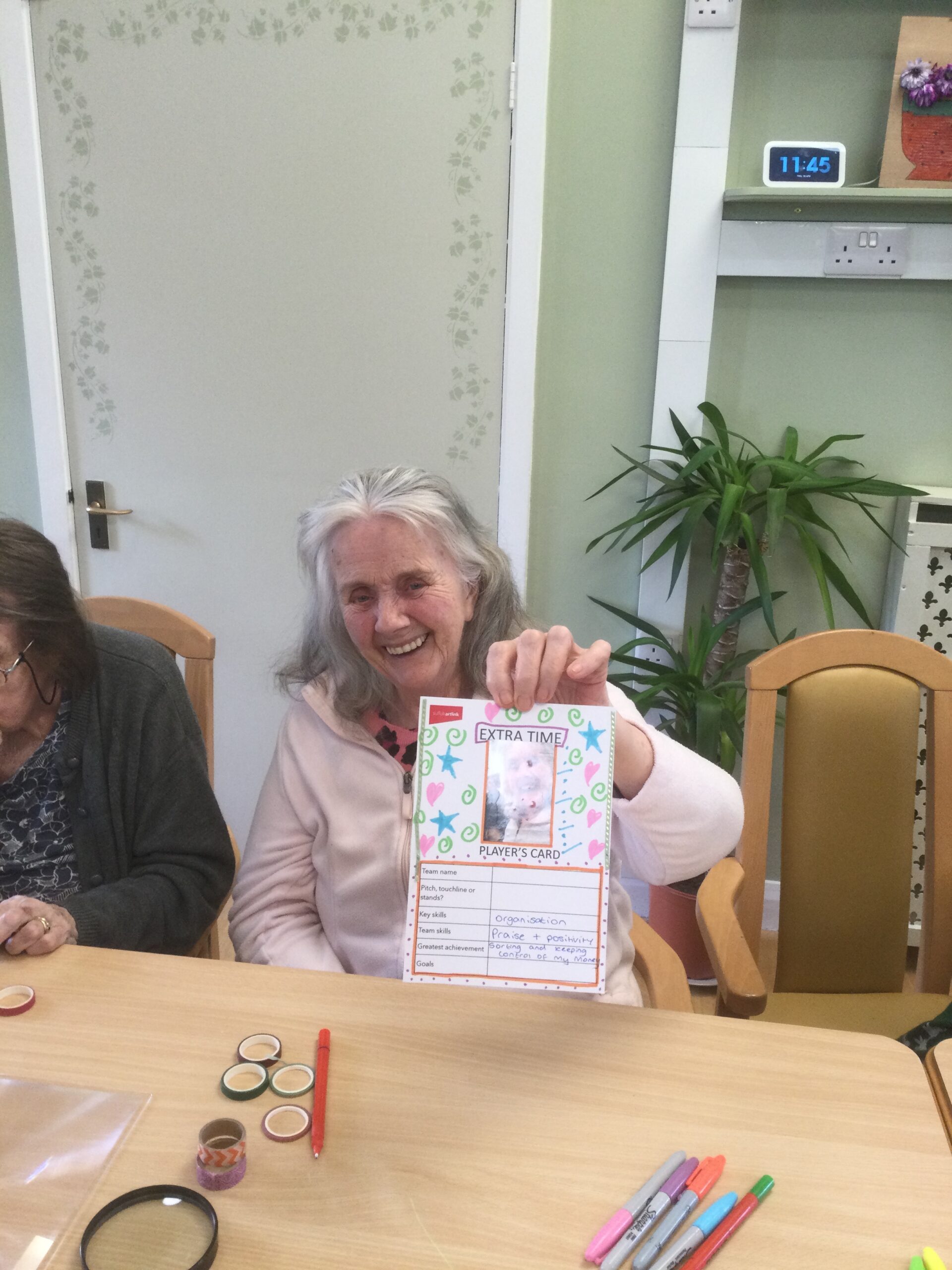 A woman smiles broadly at camera. She's holding a decorated card, covered with blue stars and pink hearts. There are pens and coloured tapes on the table in front of her.