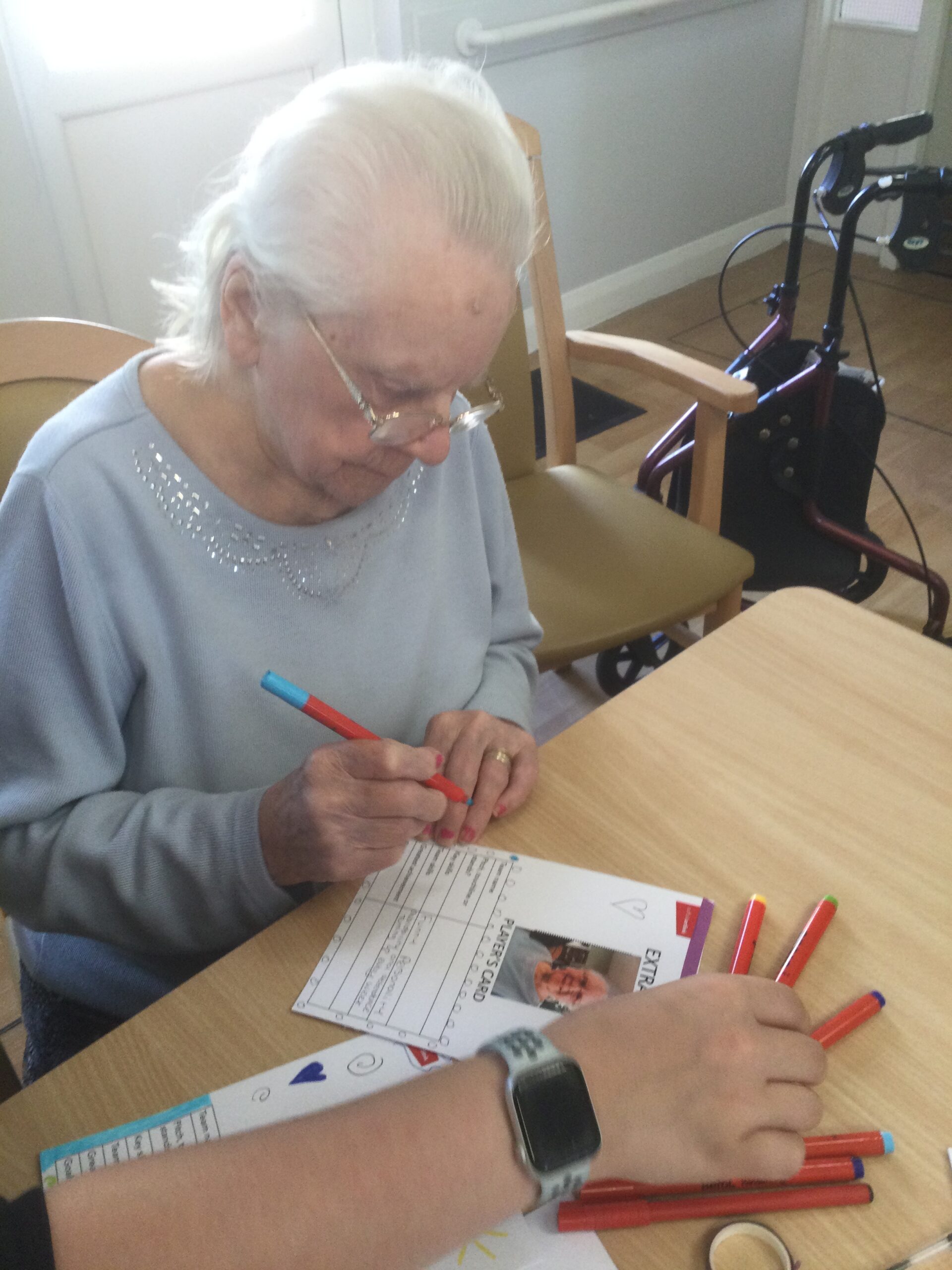 A woman in a light blue jumper with silver stitching around the neck is holding a pen, about to write something on the card in front of her. Another hand stretches in front of her, picking up a coloured pen from the table.