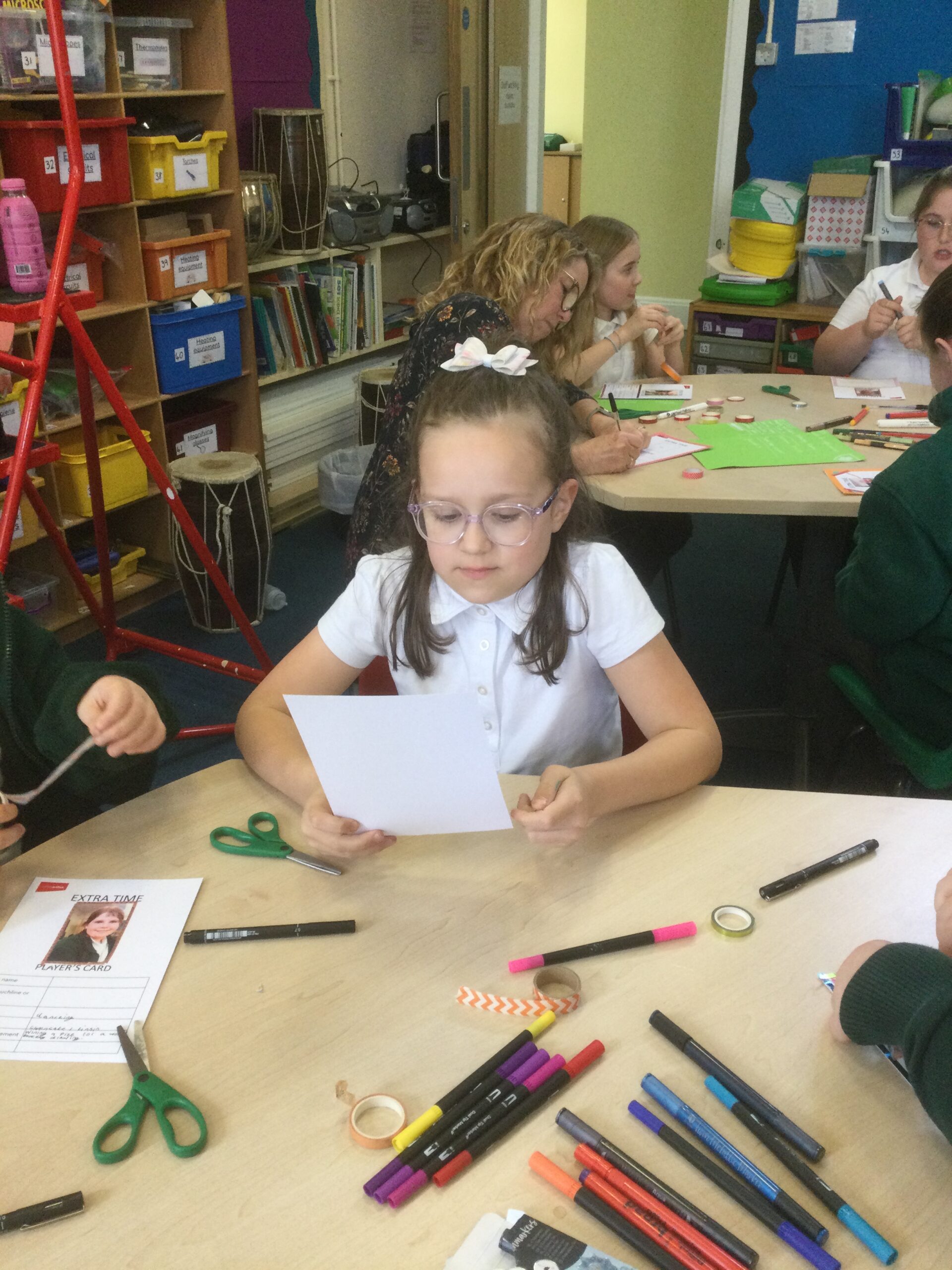 A child sits reading something from a card whilst others around her use coloured pens and tape to decorate their cards