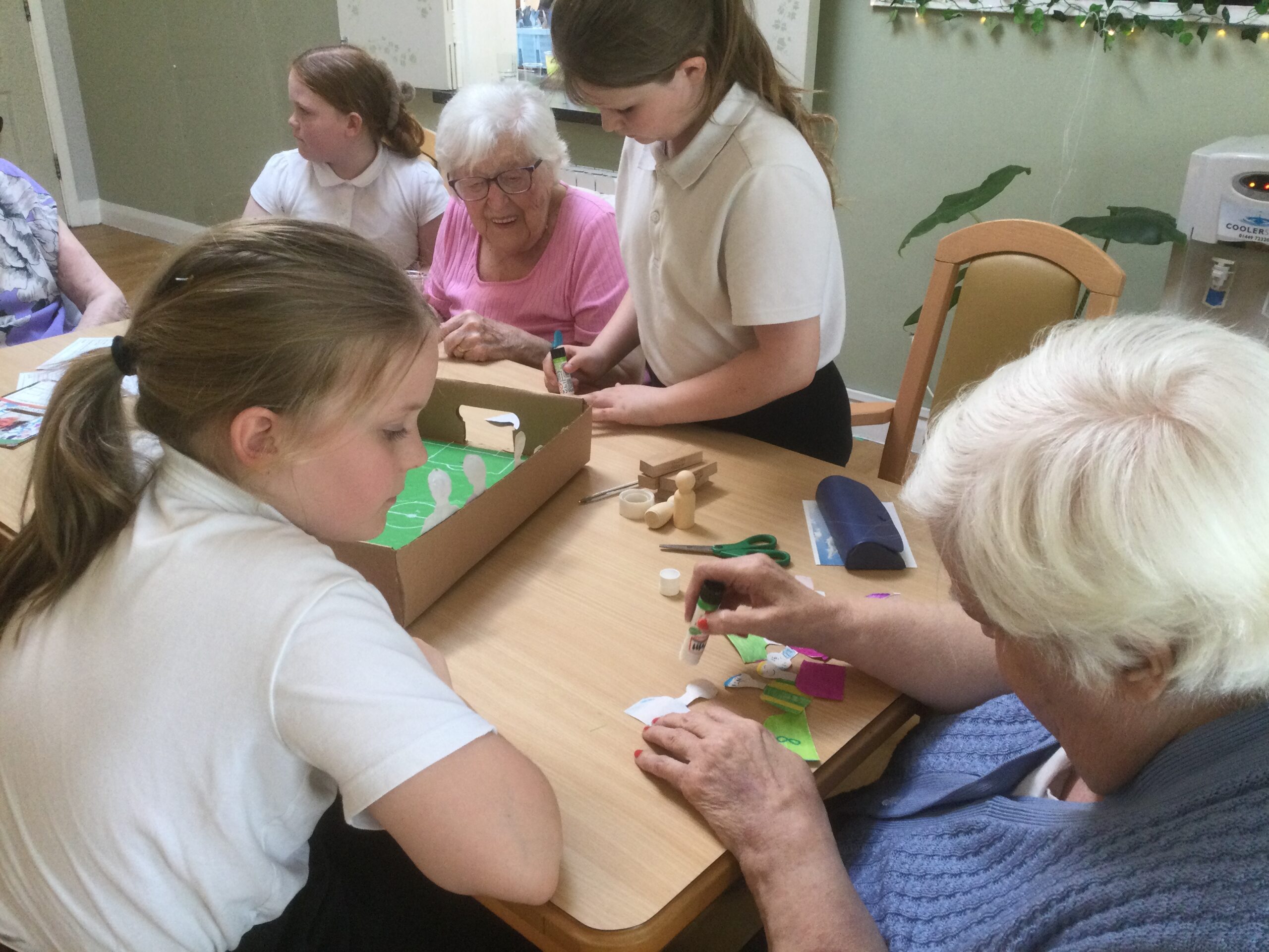 A child looks on, as an adult applies glue to a paper figure. On the other side of the table more children and adults are also gluing figures