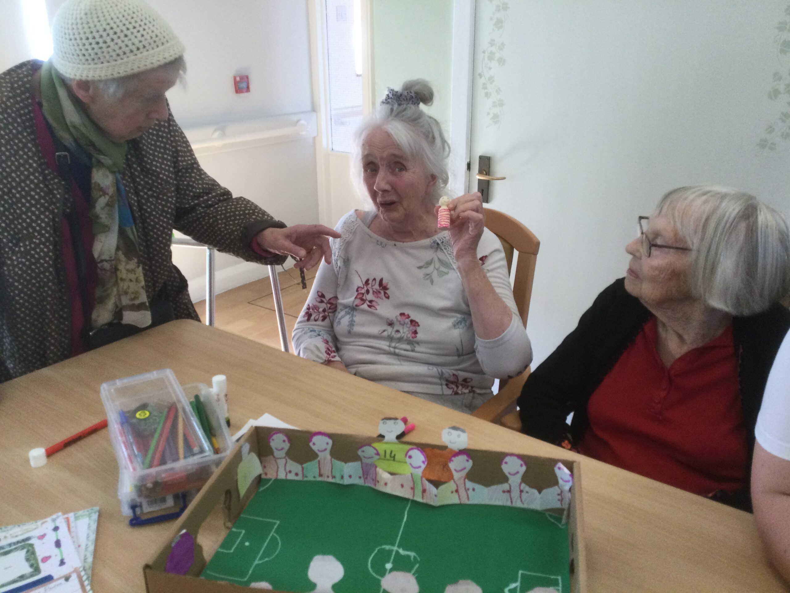 Three elderly ladies in a row, one standing two sitting, one of whom holds up a small wooden figure