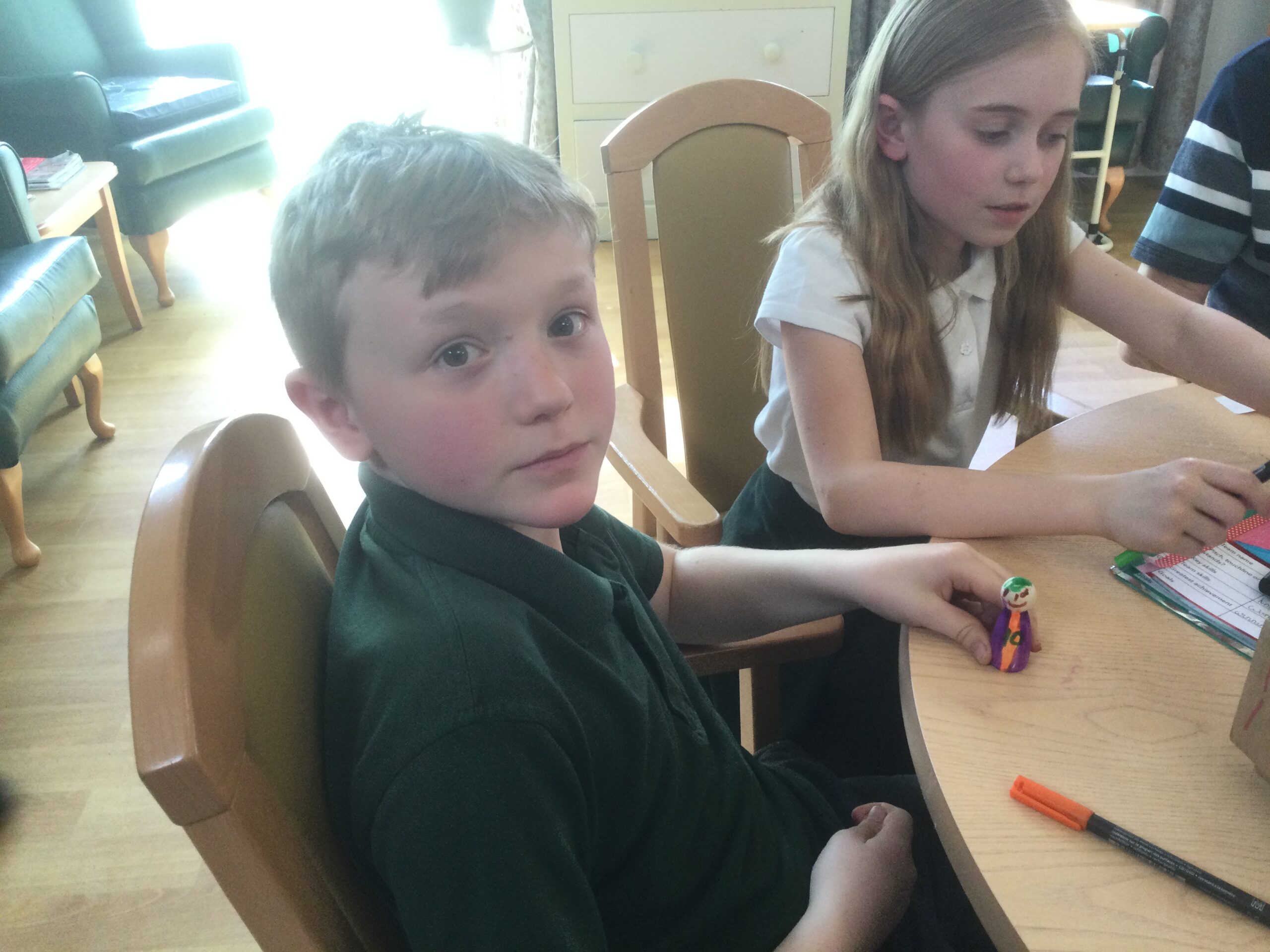 Two school children seated at a table, in front of one is a wooden figure coloured in purple and orange