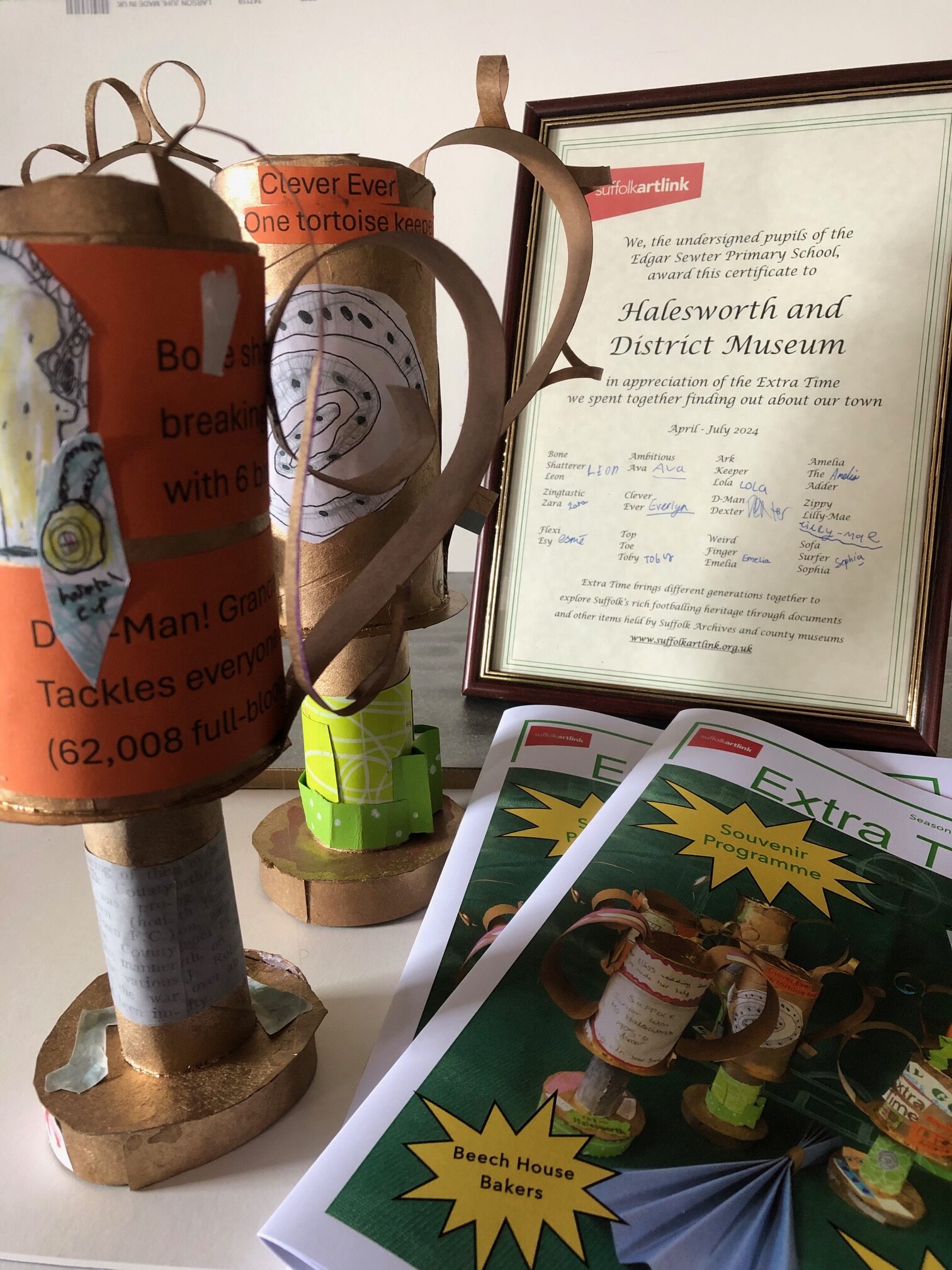 A framed certificate to the Halesworth and District Museum that reads We, the undersigned pupils of the Edgar Sewter Primary School award this certificate to Halesworth and District Museum in appreciation of the Extra Time we spent together finding out about our town - followed by several signatures. Also in the picture are two hand-crafted trophies and two Extra Time publications