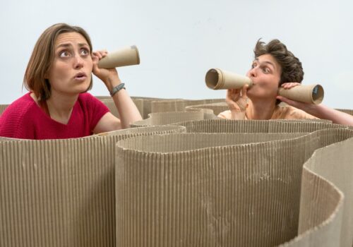 2 people amongst winding cardboard. 1 holding tube a tube like a bugle and both holding tubes like ear trumpets.