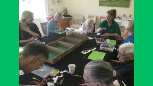 A group of people around a table strewn with shallow cardboard boxes lined with green paper, pens and sheets of paper