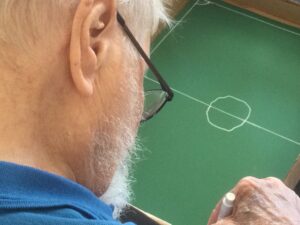 An overhead close-up of a gentleman, wearing a blue top and glasses, drawing with a white pen on green paper clearly marking out a football pitch