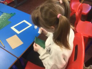 A child with hair tied in bunches with pink bands is applying glue to a piece of green paper. On the table in front of there is a blank brown postcard and a cream coloured picture frame