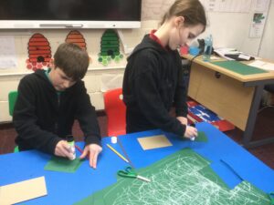 Two children applying glue to green card. On the blue table in front of them is a large sheet of green card covered in white swirls, a pair of green handled scissors and some pencils