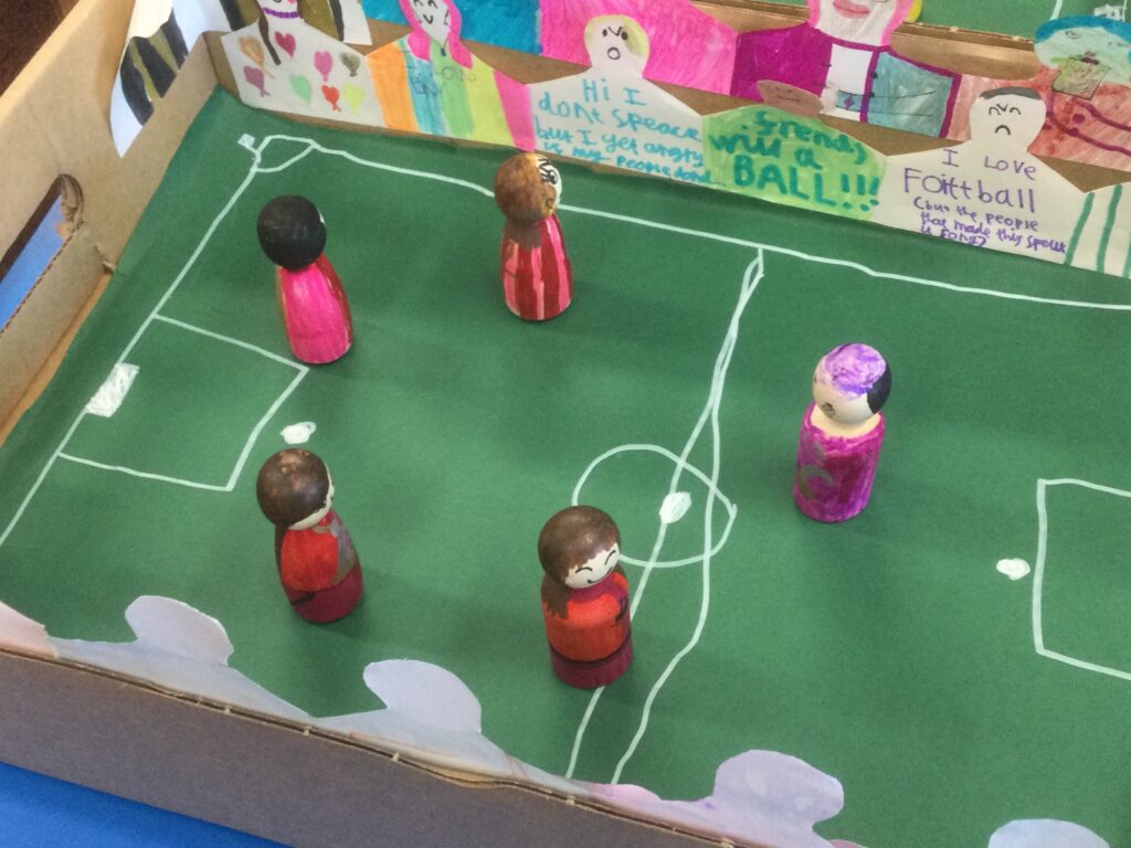 Five wooden football figures in a cardboard box stadium with rows of hand drawn spectators looking on