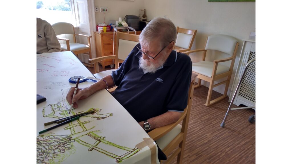 A gentleman in a blue short sleeved shirt sits at a table drawing on a long piece of lining paper taped to the table. His picture is of a tree and a paddock fence with a horse grazing