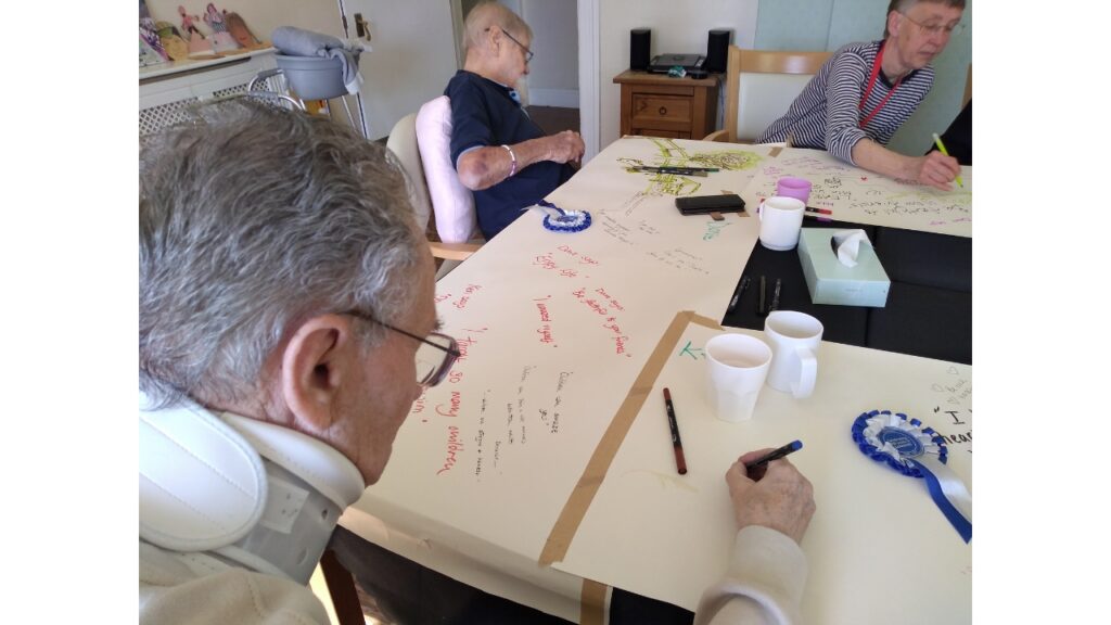 3 people seated at a table onto which is taped long strips of lining paper that they're writing on. There are cups and pens on the table as well as two blue and white rosettes