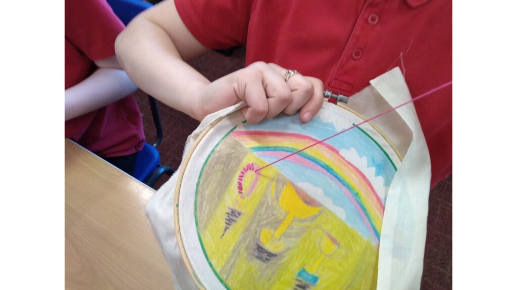 There's a colourful fabric picture in an embroidery hoop, held by someone's hand in the other hands she's pulling through a length of pink yarn