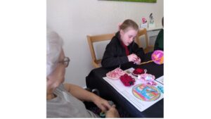 A school girl concentrates intently on a piece of crochet in her hand, a large ball of colourful wool to her let, whilst an adult looks on. In front of her is an array of crocheted items