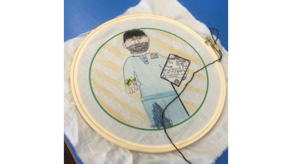 An embroidered figure, with lime green stitched irises and holding a copy of The Hooter. He has stitched black hair and a blue stitched motif on his trousers. A black thread runs across the fabric