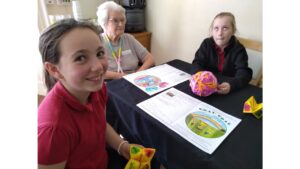 A school girl in a red top beams to the camera, she holds a yellow fortune teller in her hands. Across the table another child holds a large ball of colourful wool and rolls her eyes towards the ceiling. A resident seated between them, wearing a crochet strap around her neck, looks on smiling