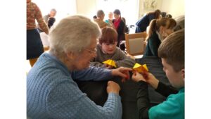 A roomful of people seated at tables covered with black cloths. In the forefront is a woman with silver hair, wearing glasses and a blue textured jumper, seated with two boys each of whom has a yellow fortune tell in his hand