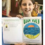 A child holds up her art work and beams at the camera, The picture is a beach scene with sun rising and blue sky and sandy beach
