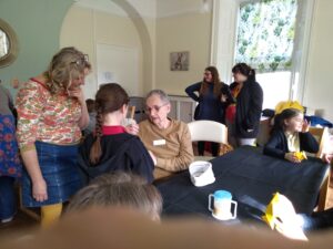 A group of people, adults and children, some seated at tables some standing. A woman on the left wearing a denim skirt and orange and sage coloured jumper with her glasses pushed up into her blonde hair, is looking down towards a seated gentleman who is taking notice of something a child has shown him