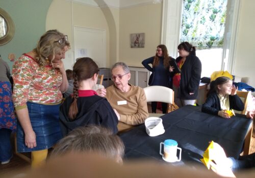 A group of people, adults and children, some seated at tables some standing. A woman on the left wearing a denim skirt and orange and sage coloured jumper with her glasses pushed up into her blonde hair, is looking down towards a seated gentleman who is taking notice of something a child has shown him