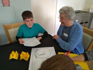 A woman in a dark floral top and blue cardigan is seated next to a boy wearing a green short-sleeved collared top over a black long sleeved T-shirt. They are seated at a table covered in a black cloth and there are two bright yellow fortune tellers and some worksheets on the table - there is also someone else's arm, with red pen in hand, in the corner of the picture