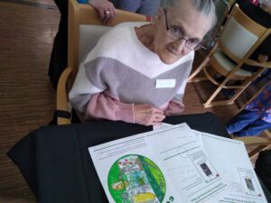 A woman seated at a table with a colourful worksheet in front of her. She is wearing a white, grey and pink jumper and has her hair swept back from her face. Someone wearing a bright ring on their finger is leaning on the back of the seated woman's chair