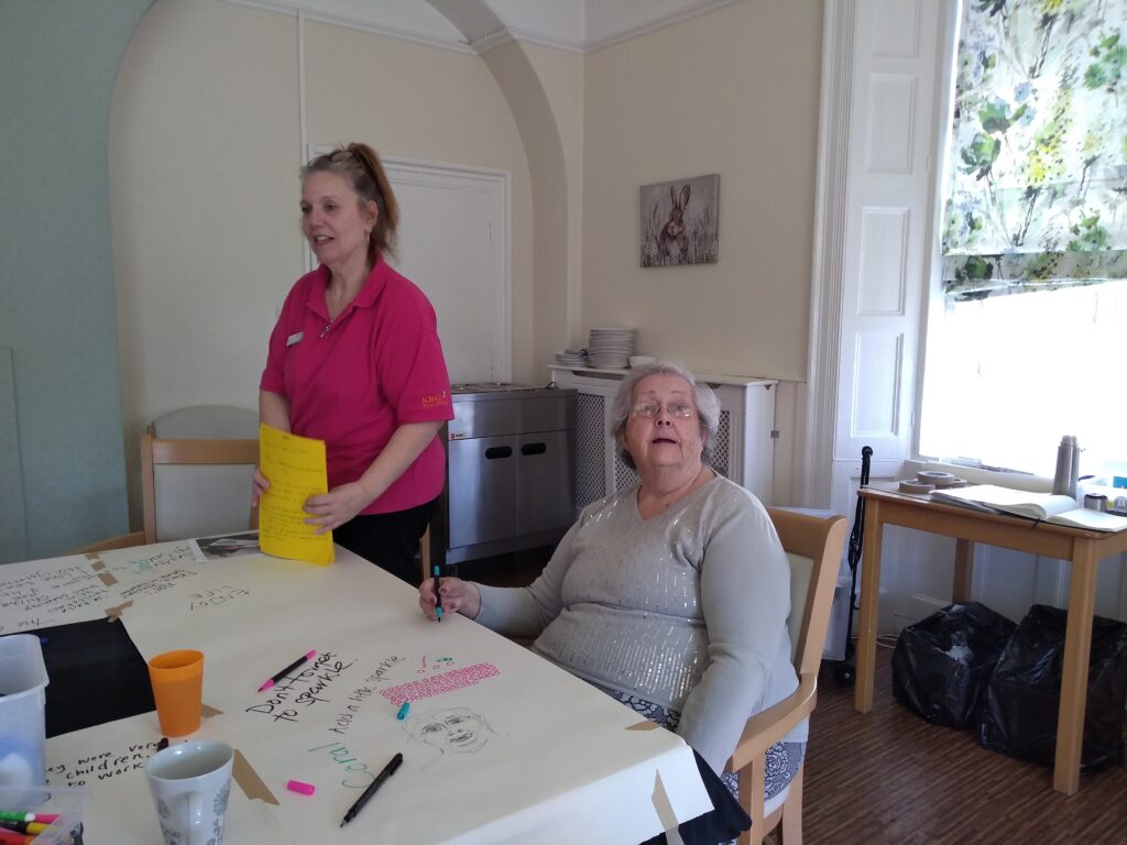 A woman wearing a grey top with silver sequins down the front is seated at a table holding a pen in her right hand. Beside stands another woman wearing a cerise short sleeved top and with her hair tied back in a pony tail. She holds a stack of yellow papers vertically in her hands. The table has an array of pens, papers and mugs and cups