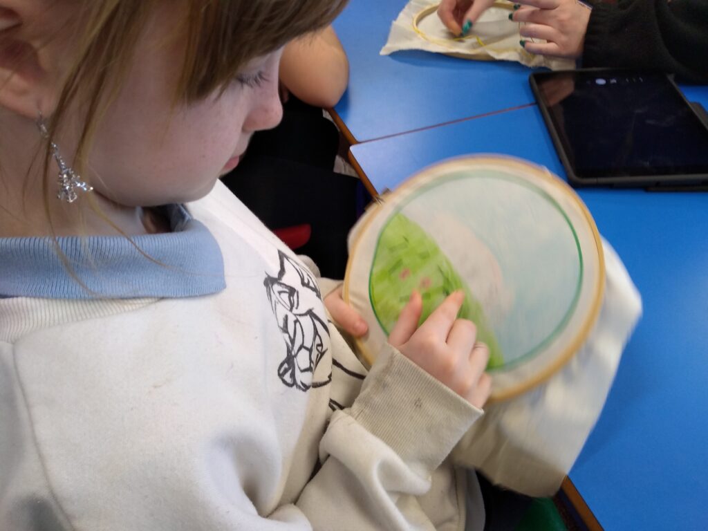 An overhead view of a child holding a fabric picture in an embroidery hoop pointing to an element in the images. She is wearing a dangling earring that looks like the Eiffel Tower