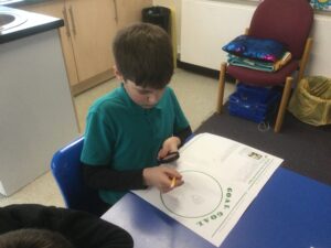 A school boy holds a magnifying glass in one hand and a yellow pencil in the other. He is looking through the glass as he writes something on a worksheet in front of him. He is working at a blue-topped table and to his right is another chair with colourful cushions