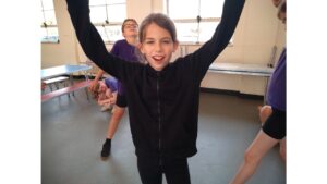 A child faces camera, her arms stretched upwards and a cheering look on her face. A young lad with glasses stands behind her with his chin up and arms outstretched at his sides