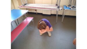 A child crouches on the floor, arms hugged around her knees and head tucked in. Around her are long tables and benches, some stacked on top of each other