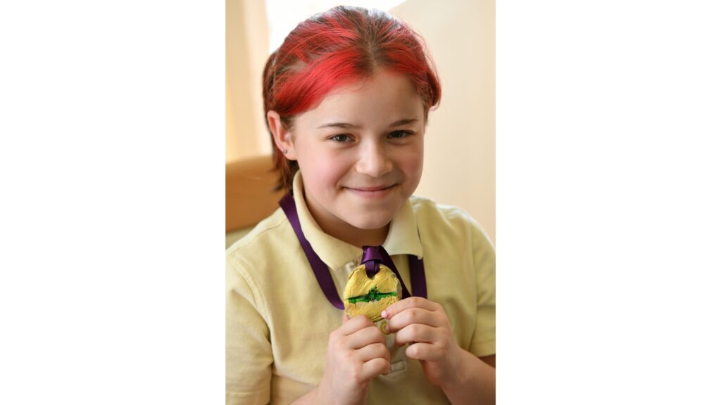 A young girl wearing a yellow top and with red highlights in her fringe holds up a gold medal with a green line through the centre strung on a purple ribbon