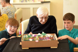 A white-haired gentleman in a dark top and wearing glasses is seated at a table covered with a black cloth. He is flanked by two school boys dressed in green tops and in front of them on the table is a shallow brown cardboard box crafted into a football pitch with colourful football fans pasted to the sides. The boy on the left is holding a long slender knitting needle with a small wooden figure attached to the end via a magnet