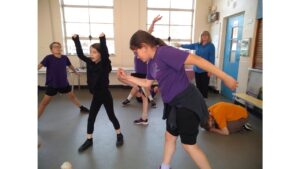 A group of children in a school hall, adopting a variety of poses with arms up, legs outstretched and crouched on the floor