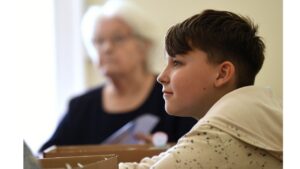 A young boy in a white and grey speckled hoodie looks directly ahead. Blurred background is a woman with white hair.
