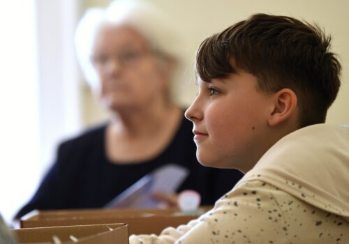 A young boy in a white and grey speckled hoodie looks directly ahead. Blurred background is a woman with white hair.