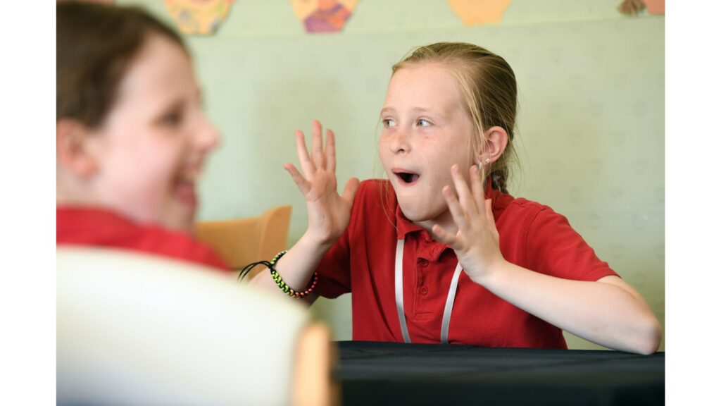 A young girl, mouth agape and hands in the air. Blurred foreground another schoolgirl laughing