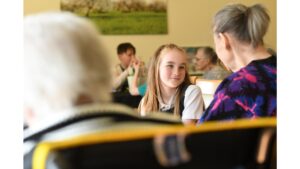 A school girl, wearing a white top and her long hair swept back and hanging down either side of her face, looks warmly towards a person seated to her left. Blurred people in immediate fore- and back ground