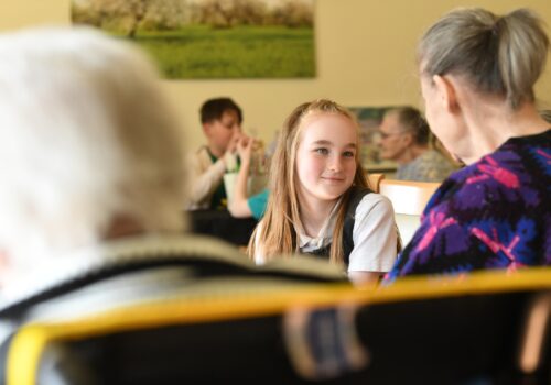 A school girl, wearing a white top and her long hair swept back and hanging down either side of her face, looks warmly towards a person seated to her left. Blurred people in immediate fore- and back ground