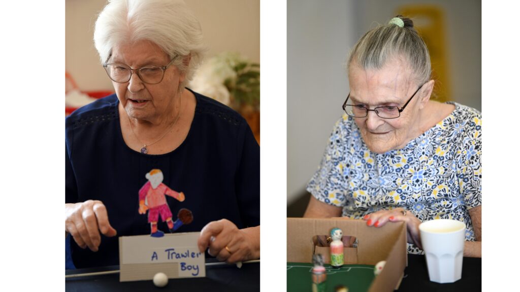 Two images - left a white haired woman wearing glasses holds a cardboard football figure A Trawler Boy and a small white ball in front. Picture right - a woman with hair swept back in a band and wearing a blue and yellow floral top plays a table top football game.
