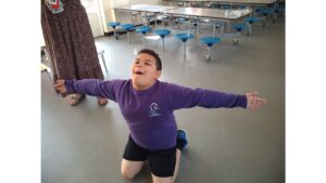 A child in purple top and black shorts with his arms outstretched demonstrating a knee slide on the hall floor. Behind him are tables with integral circular blue stools and a person in a brown skirt and sandals to the left