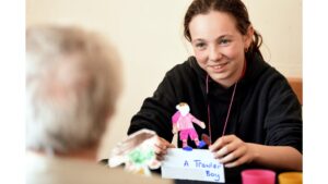 A school girl in a dark top, her hair swept away from her face, and holding a cardboard football figure in her hands on the table A Trawler Boy looks across the table towards another person