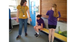 A child sits on a wooden school bench with another child standing to her left. An adult wearing jeans and ginger top and green lanyard round her neck looks towards the seated child and holds her hand towards her