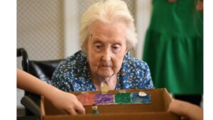 An elderly lady dressed in a blue, white and yellow patterned dress, looks intently at a game of table football.