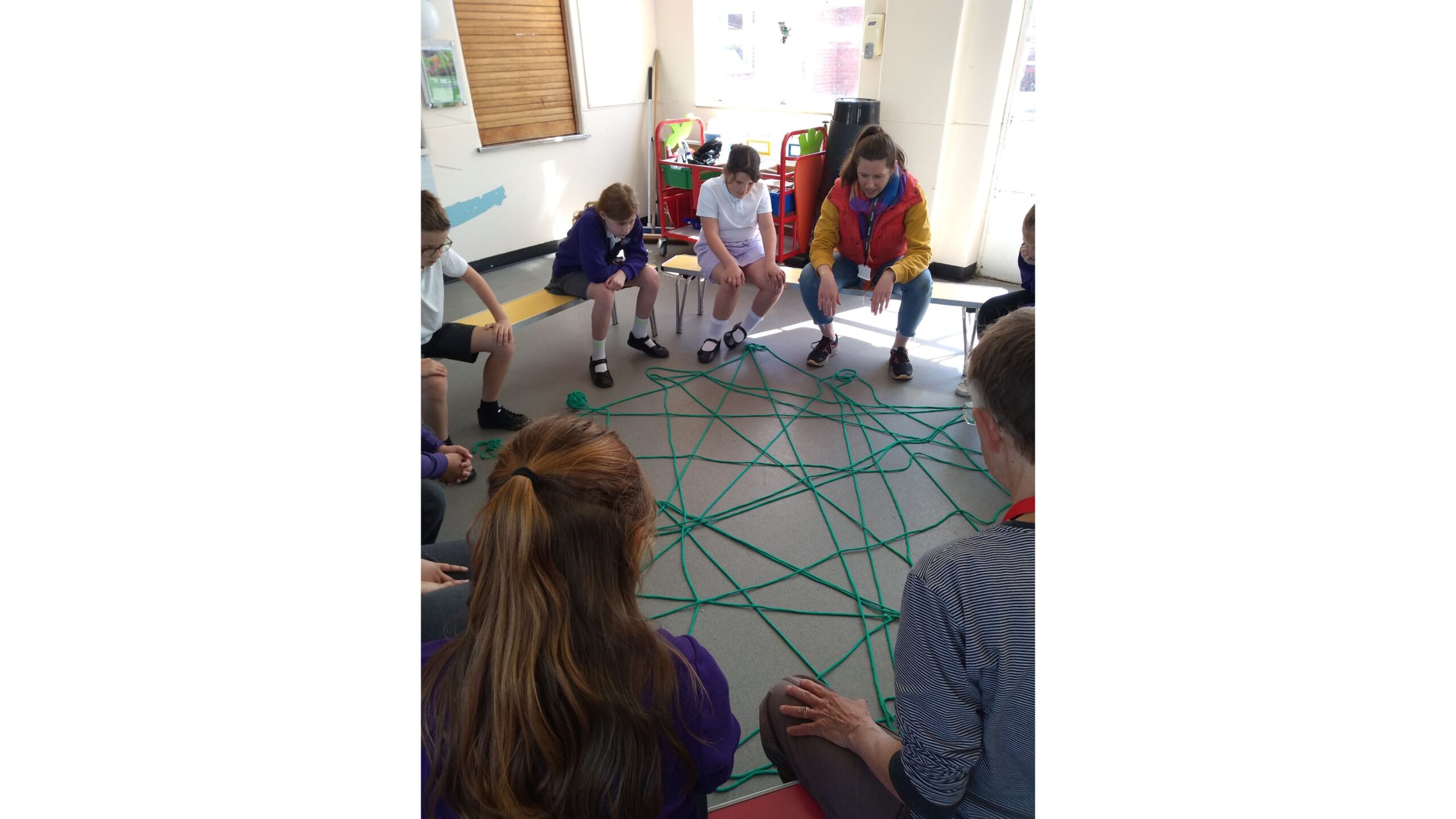 A group of children and adults seated on school benches. On the floor in front of them is a large 'net' of green wool, that they have created by throwing a ball of wool back and forth to one another