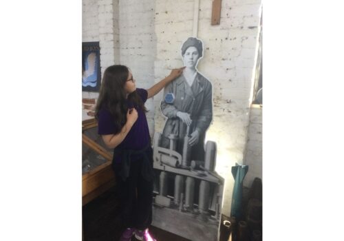 A school girl with long dark hair and wearing dark-rimmed glasses points to a life-sized figure of a WWI munitionette propped up against a white-washed brick wall