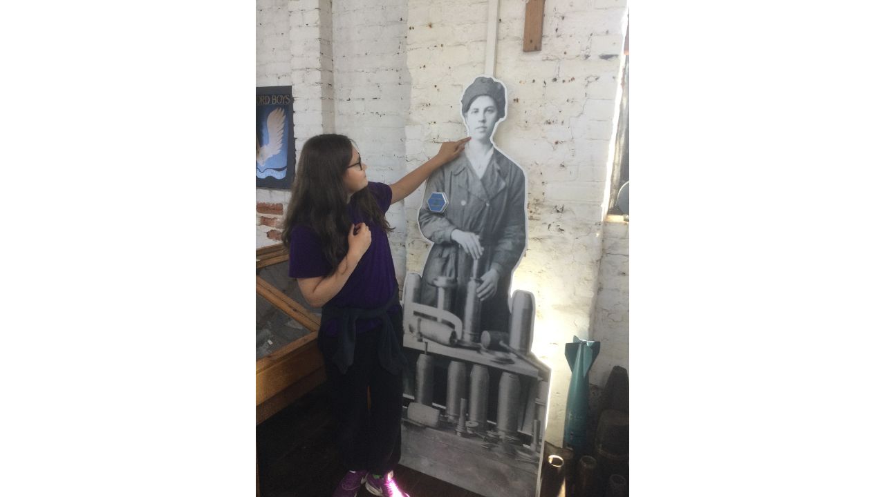A school girl with long dark hair and wearing dark-rimmed glasses points to a life-sized figure of a WWI munitionette propped up against a white-washed brick wall