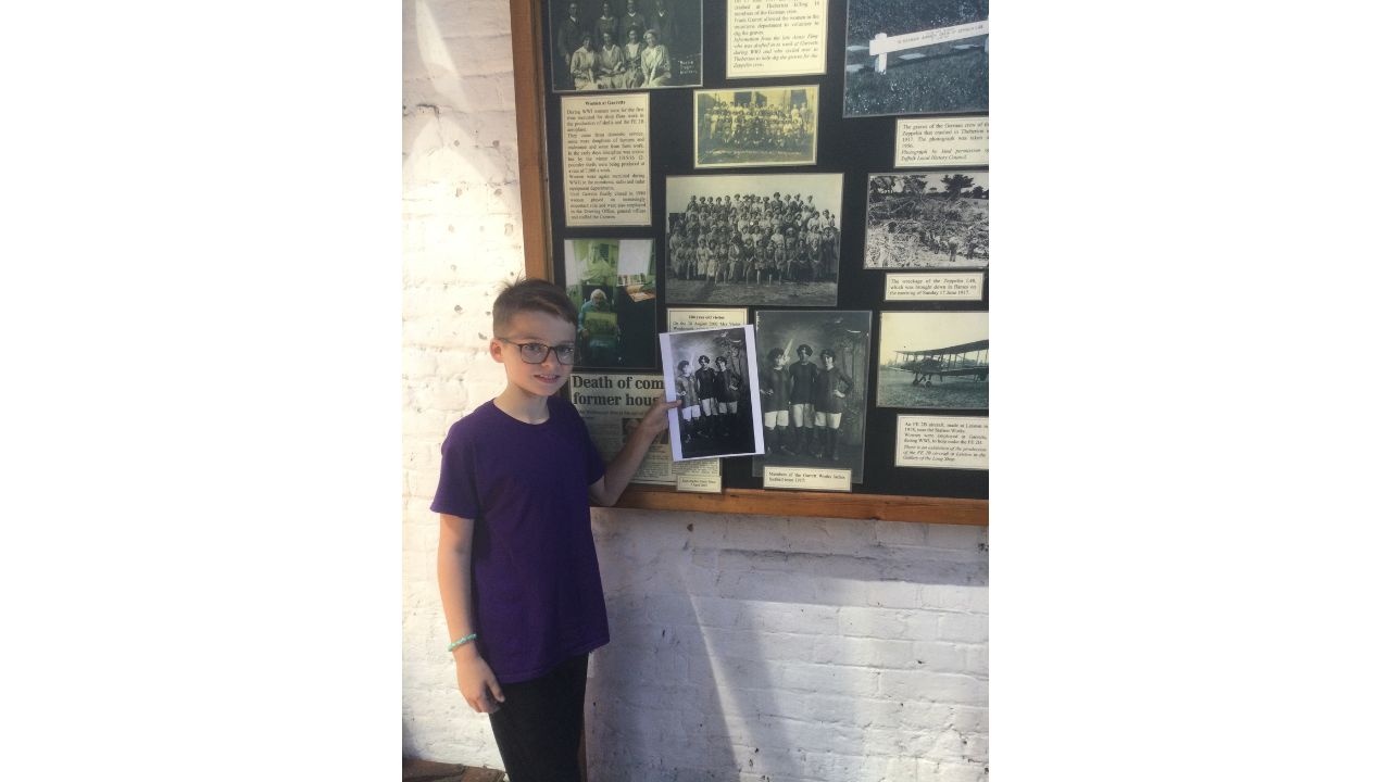 A school boy in purple T-shirt and black trousers stands in front of a large wall display of monochrome photographs and text. He holds a photograph in his hand, placing it beside an identical picture on the display board