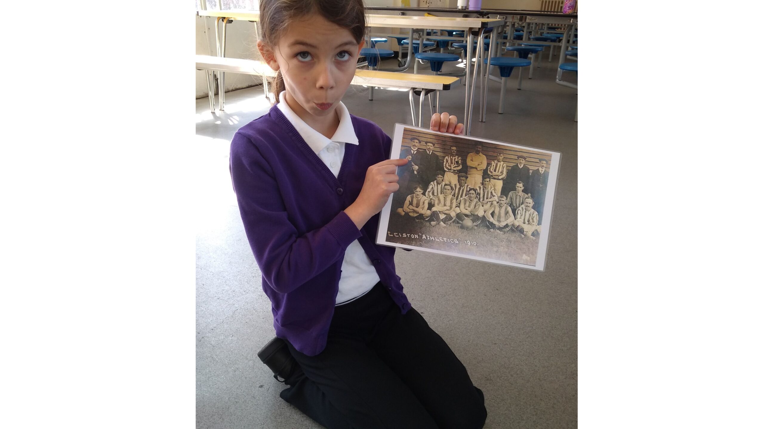 A school girl kneels on the floor, holding an old sepia photograph of a football team. She indicates on of the people in the picture and is pulling a long face, as if impersonating the individual