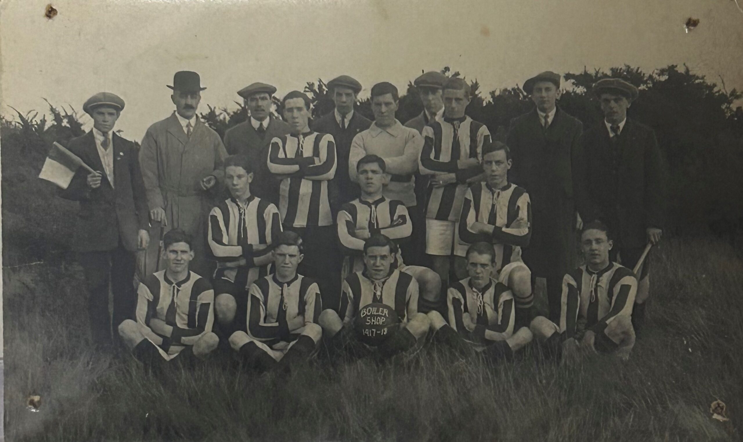 A monochrome photograph of a football team, in the centre a player holding the football with the words Boiler Shop 1917 - 1918 on it