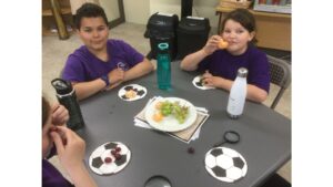 Children seated at a square gray table with a plate of green grapes and satsuma segments in the middle and football napkins in front of them